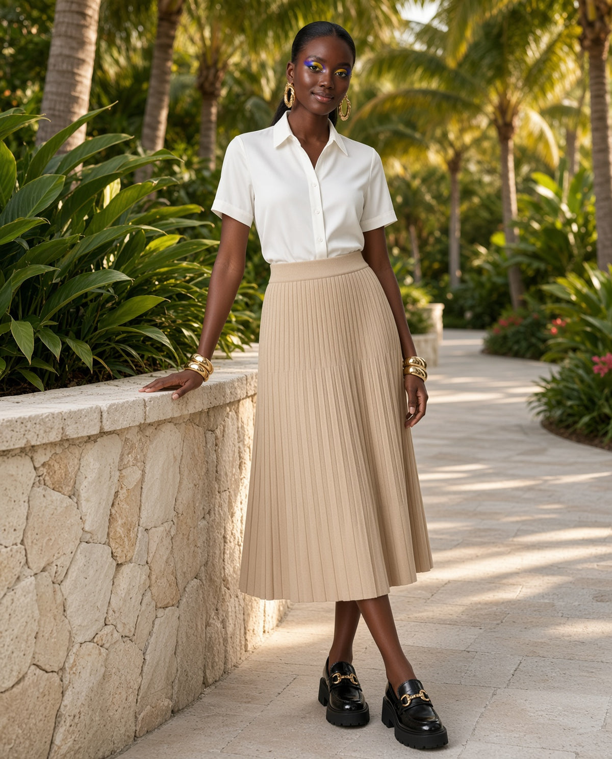 Model wearing the Chelsea Pleated Midi Skirt in tan on a palm-lined garden path